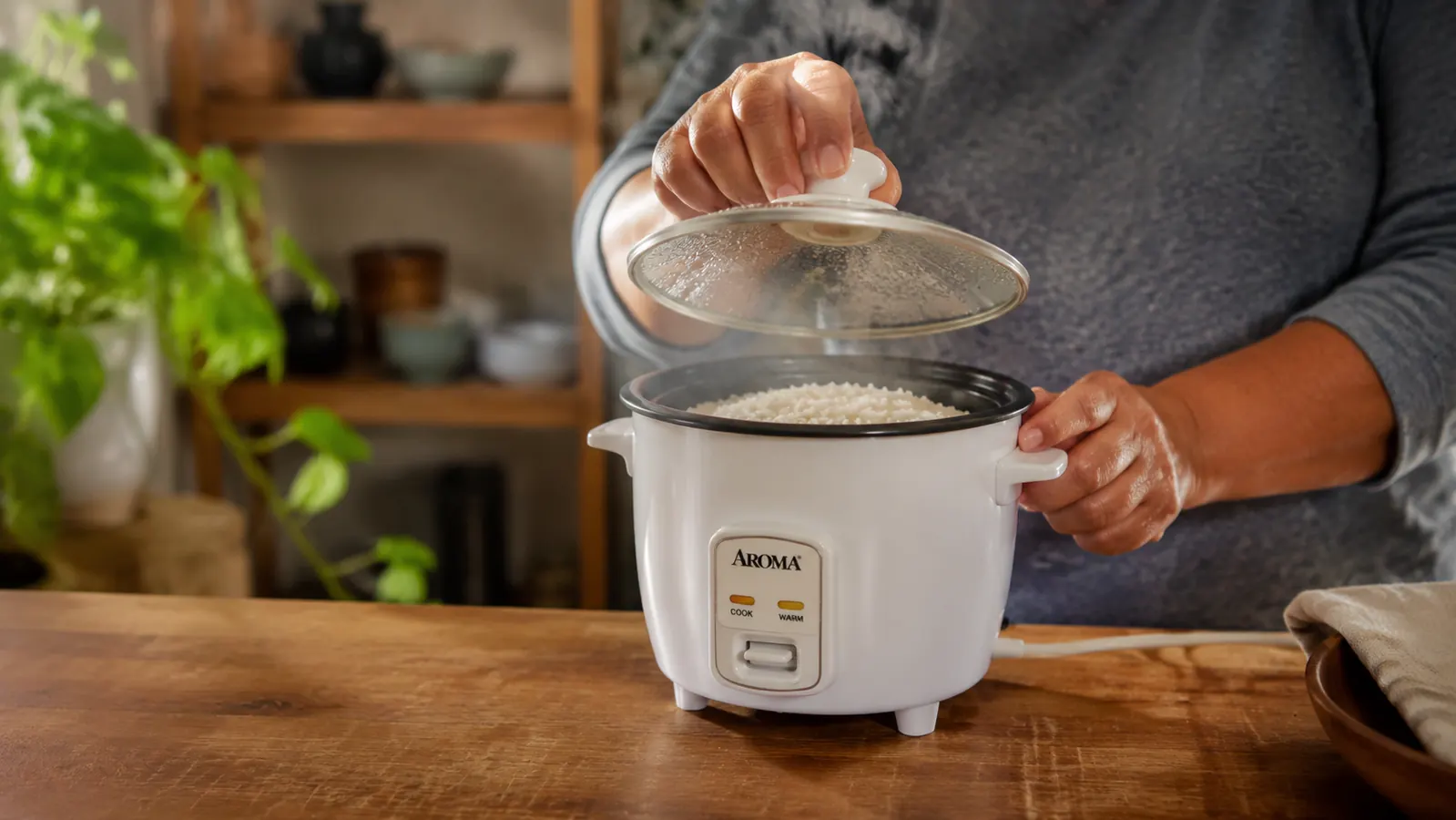Hands gently lifting the lid of an Aroma rice cooker to check on steaming rice inside